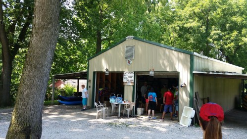 A green and beige shed surrounded by trees, with people gathered outside and kayaks visible nearby.