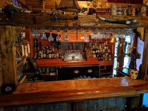 A rustic bar with wooden decor, shelves lined with various bottles, and hanging glasses in a cozy setting.