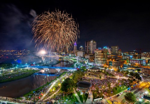 A vibrant fireworks display over a city skyline, with a large crowd gathered by a riverbank at night.
