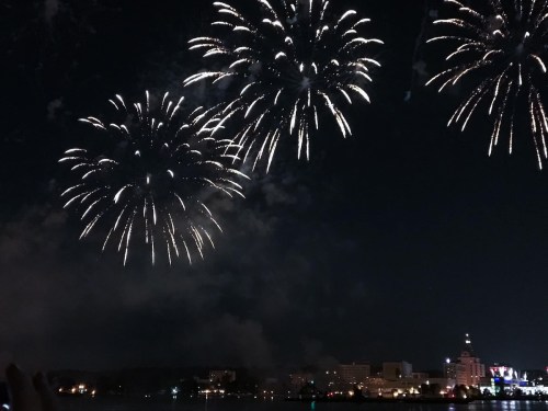 Colorful fireworks illuminate the night sky above a city skyline, reflecting on the water below.