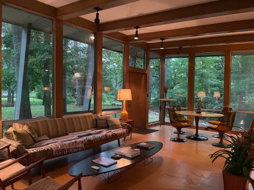 A cozy, modern living room with large windows overlooking a green landscape, featuring a sofa, chairs, and a coffee table.