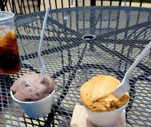 Two cups of ice cream, one chocolate and one caramel, sit on a black metal table next to a drink.