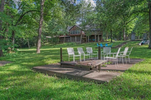 A grassy area with a fire pit surrounded by white chairs, with a wooden house and trees in the background.