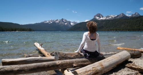 A woman wearing a white shirt sits on a fallen log, looking toward Redfish Lake in Idaho.