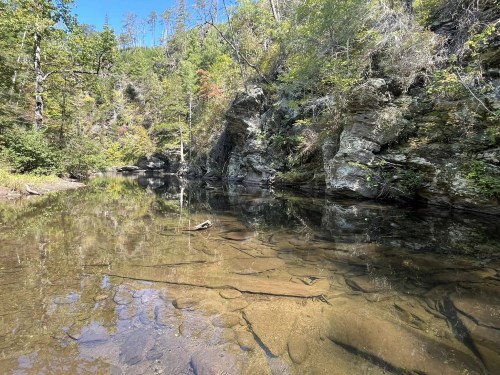A serene river scene with clear water reflecting rocky banks and lush greenery under a bright blue sky.