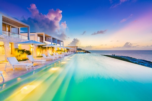A serene infinity pool at sunset, surrounded by lounge chairs and ocean views, with colorful clouds in the sky.