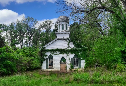 Abandoned white building with a dome, surrounded by overgrown greenery and trees under a blue sky.