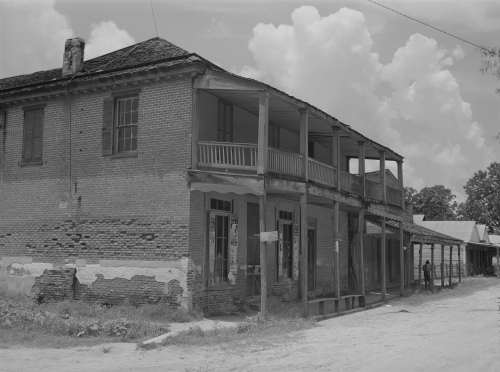 Black and white photo of a weathered brick building with a porch, surrounded by a dirt road and cloudy sky.