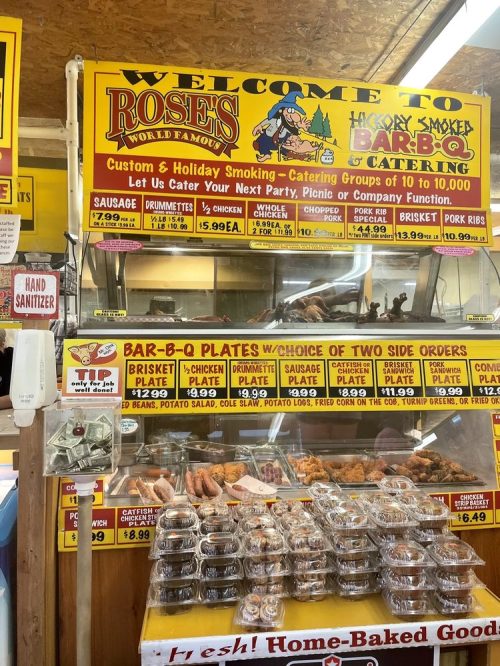 Colorful sign for Rose's BBQ, showcasing menu items like brisket, sausage, and catering options, with a hand sanitizer station.