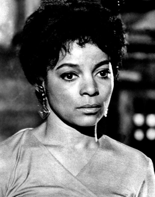 A black and white portrait of a woman with short hair, wearing a simple dress and earrings, looking thoughtfully off-camera.