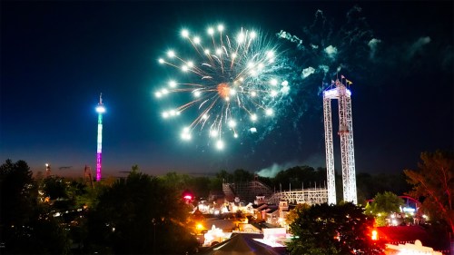 Colorful fireworks illuminate the night sky above a vibrant amusement park, with rides and trees in the foreground.