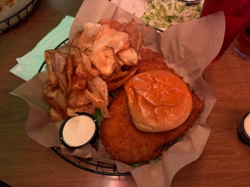 A basket containing a fried chicken sandwich with lettuce, a bun, and a side of crispy potato chips.