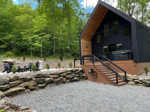 Modern black and wood cabin surrounded by greenery, featuring a gravel area, stone wall, and outdoor seating.