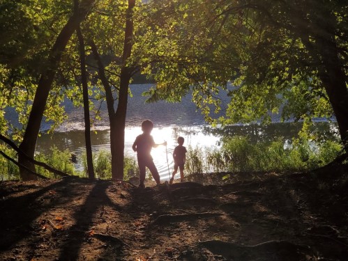 Silhouettes of two children playing by a river, surrounded by trees and sunlight reflecting on the water.