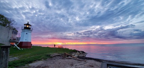 A lighthouse stands by the shore at sunset, with vibrant clouds reflecting on the calm water.