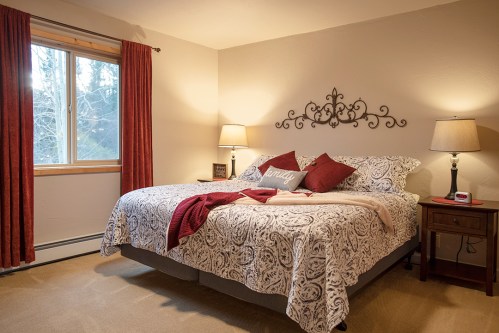 Cozy bedroom with a patterned bedspread, red curtains, and lamps beside a window overlooking trees.
