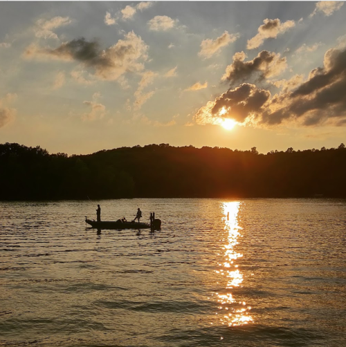 Silhouette of a boat with people on a calm lake at sunset, with golden reflections on the water and clouds in the sky.