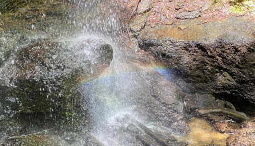 A close-up of a waterfall cascading over rocks, creating mist and a faint rainbow in the sunlight.