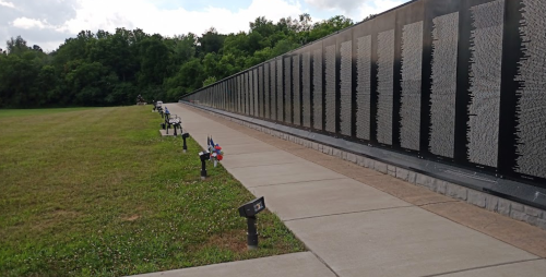 A memorial wall with names, surrounded by grass and small flags, under a cloudy sky.