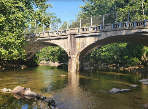 A stone bridge arches over a calm river, surrounded by lush greenery and rocky riverbanks.