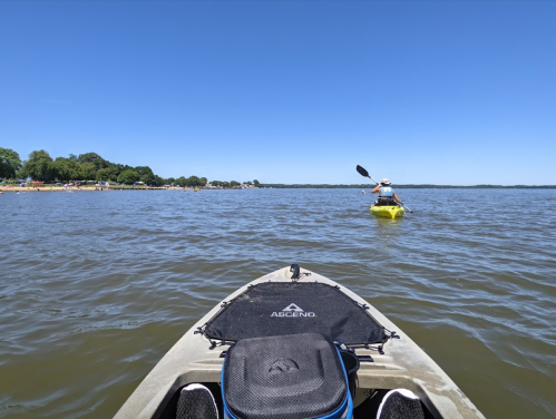 A view from a kayak on a calm lake, with another kayaker paddling in the distance under a clear blue sky.