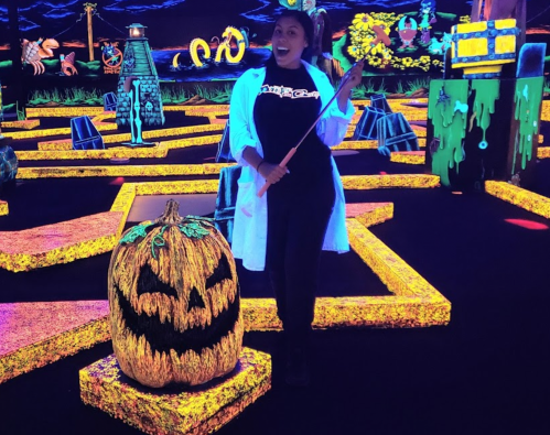 A person in a lab coat poses with a golf club next to a glowing pumpkin in a colorful mini-golf setting.