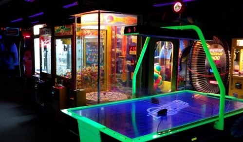 A dimly lit arcade featuring a neon air hockey table and various claw machines filled with colorful prizes.