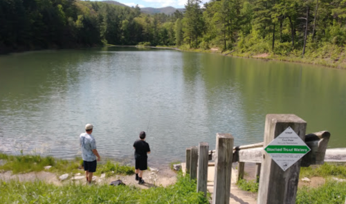 Two people fishing by a calm lake surrounded by trees, with a sign indicating the area is for stocked trout waters.