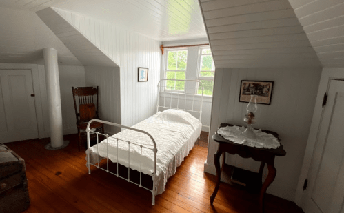 A cozy, white-walled bedroom with a single bed, wooden floor, and vintage furniture, including a rocking chair and table.
