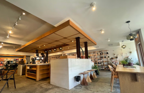 A cozy café interior featuring wooden furniture, a counter, bookshelves, and warm lighting.