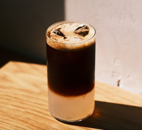 A glass of iced coffee with a frothy top, sitting on a wooden surface with soft lighting.