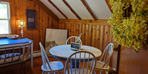 Cozy room with wooden walls, a round table, chairs, and a decorative wreath on the door. Natural light from a window.