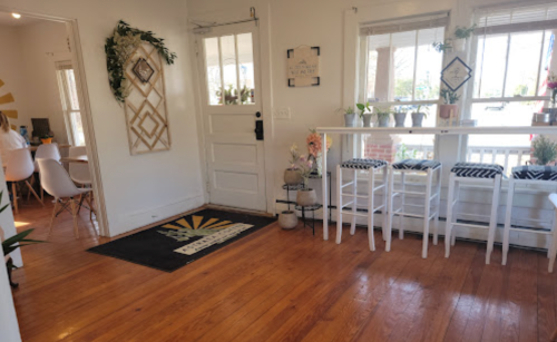 Bright interior of a cozy space with wooden floors, plants, and a small bar area with stools. Sunlight streams in through windows.