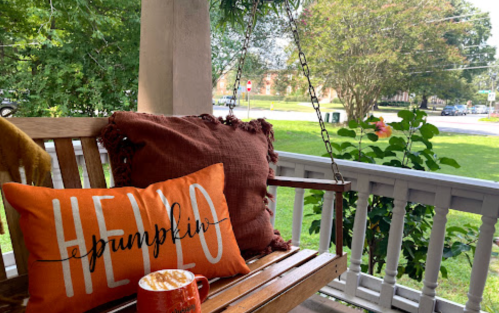 A cozy porch scene with a "Hello Pumpkin" pillow, a mug, and greenery in the background.