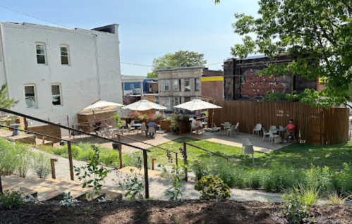 A sunny outdoor café with umbrellas, tables, and greenery, surrounded by buildings and a wooden fence.