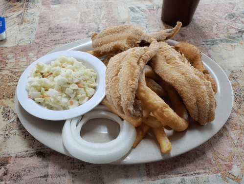 A plate of fried fish fillets with coleslaw, fries, and onion rings on a patterned tablecloth.