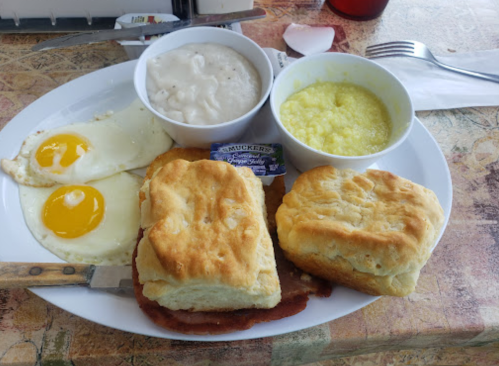 A plate with two fried eggs, biscuits, gravy, and a bowl of grits. A knife and a packet of jam are also on the plate.
