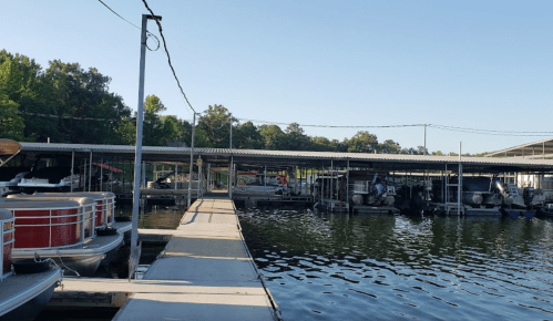 A marina with a wooden dock, lined with boats and covered slips, surrounded by trees and calm water.
