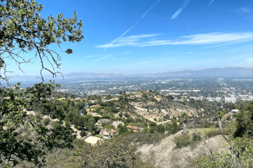 A panoramic view of a valley with houses, greenery, and a clear blue sky with wispy clouds.