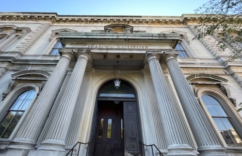 Historic building entrance with tall columns and a sign reading "Peabody Institute." Clear blue sky above.