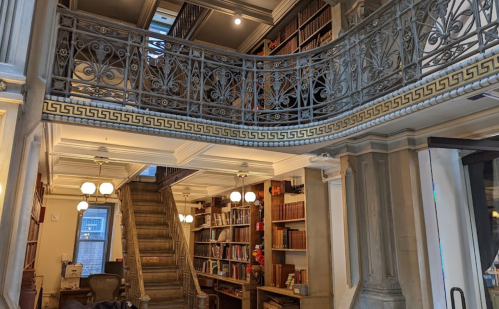 A grand library interior featuring ornate railings, bookshelves, and a staircase leading to an upper level.