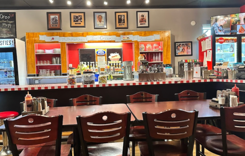A cozy diner interior with wooden tables, a counter displaying ice cream, and framed photos on the walls.
