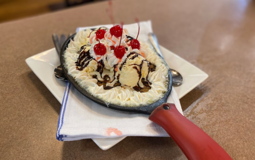 A sizzling dessert on a skillet, featuring ice cream, whipped cream, chocolate sauce, and cherries on top.