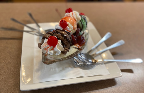 A glass dish filled with ice cream scoops, whipped cream, and cherries, surrounded by several spoons on a plate.