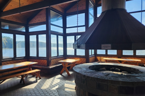 Cozy lakeside cabin interior with large windows, wooden tables, and a stone fireplace, offering scenic views.