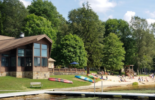 A lakeside scene with a beach, people enjoying the sun, and a wooden building surrounded by trees.