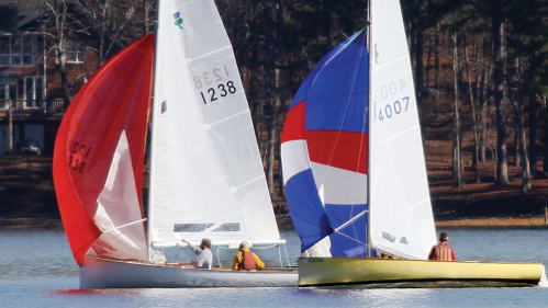 Two sailboats race on a lake, one with a red sail and the other with a blue and white sail, surrounded by trees.