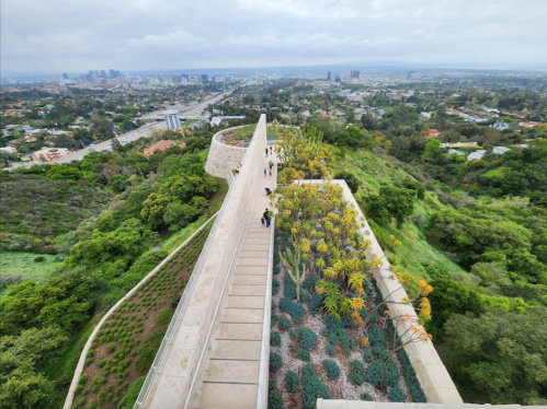 Aerial view of a landscaped walkway on a hillside, overlooking a cityscape with greenery and distant buildings.