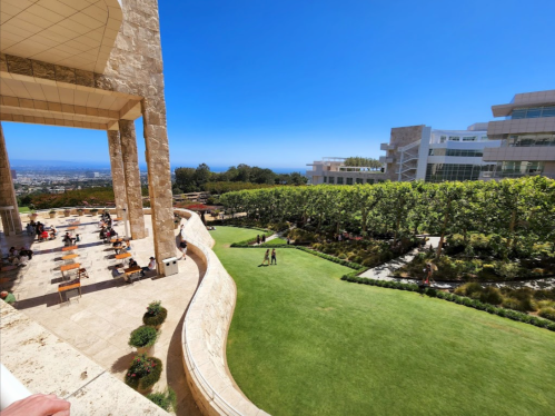 A sunny outdoor view of a landscaped area with people walking, surrounded by modern architecture and greenery.