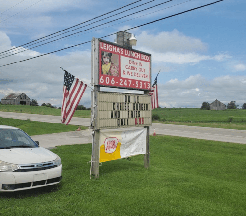 Sign for Leigha's Lunch Box advertising cheeseburgers and fries, with American flags and a rural backdrop.